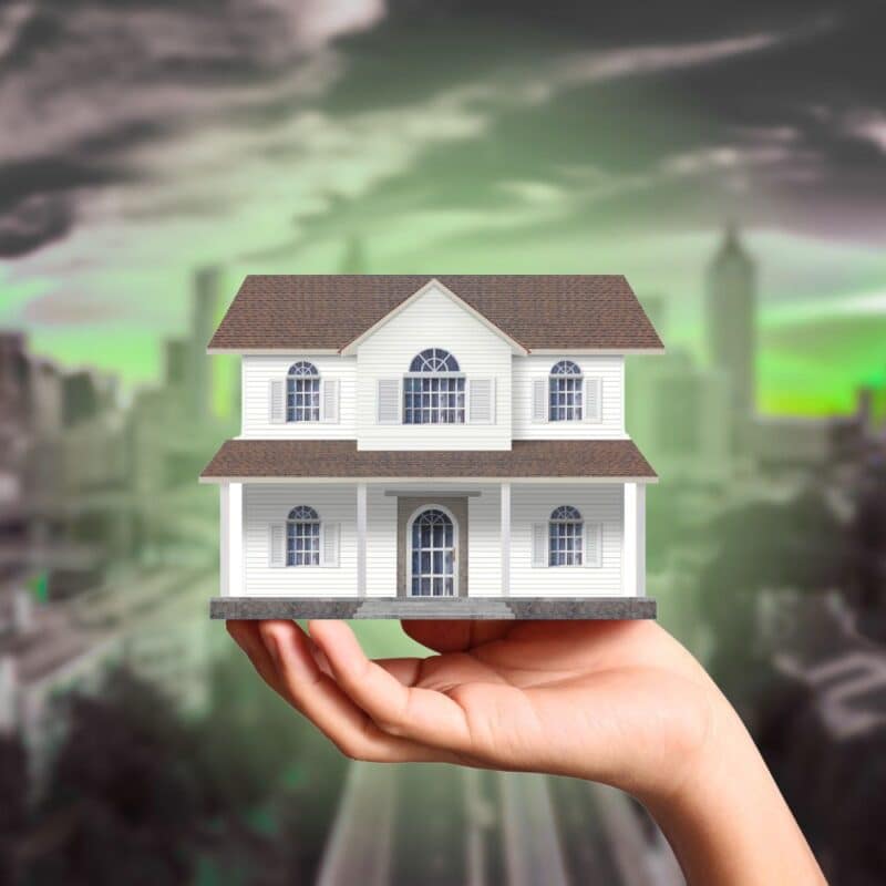 A hand holding a small model of a white two-story house against a blurred cityscape with a dark green-tinted sky.