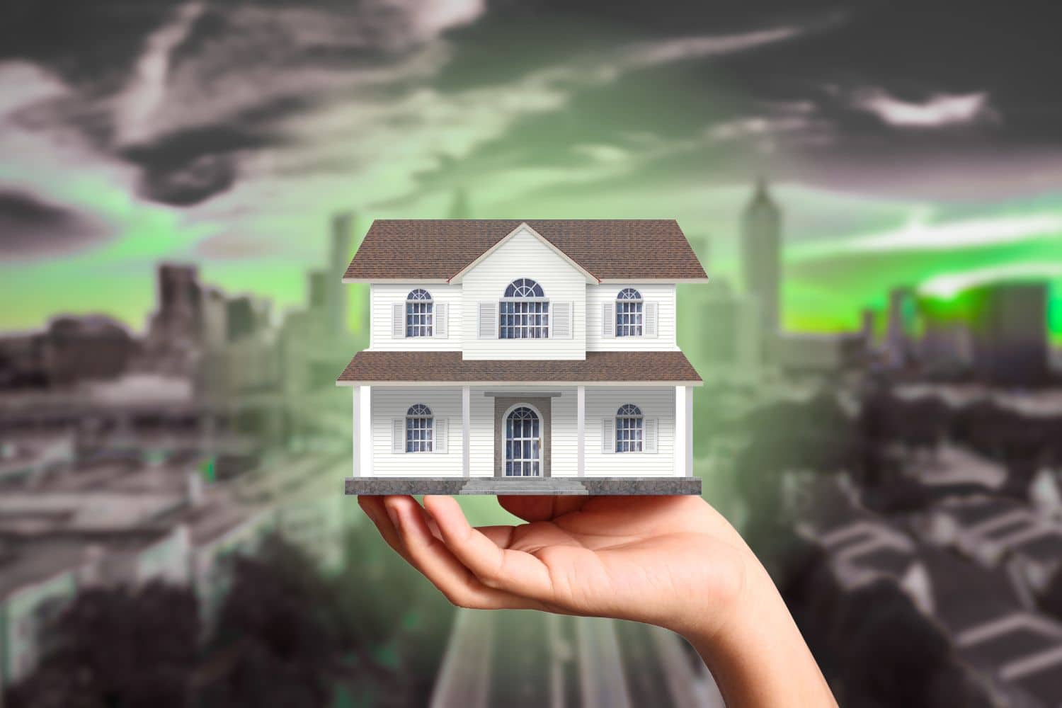 A hand holding a small model of a white two-story house against a blurred cityscape with a dark green-tinted sky.